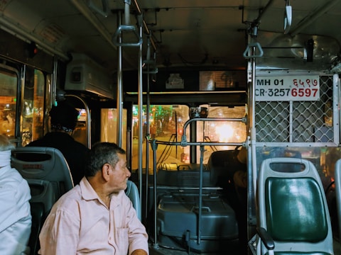 Inside view of a public bus at night with several passengers seated. A man in a light shirt sits near the front, gazing out the window. The bus interior features seats, metal bars for stability, and a partial view of the driver. External street lights create reflections on the windows.
