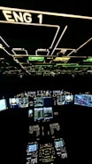 A cockpit filled with illuminated panels and displays showing various controls and instruments. The main displays are active with complex data and indicators. The environment is dark, highlighting the bright green, blue, and white lights from the instruments.