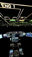 A cockpit filled with illuminated panels and displays showing various controls and instruments. The main displays are active with complex data and indicators. The environment is dark, highlighting the bright green, blue, and white lights from the instruments.