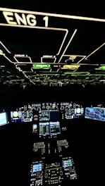 A cockpit filled with illuminated panels and displays showing various controls and instruments. The main displays are active with complex data and indicators. The environment is dark, highlighting the bright green, blue, and white lights from the instruments.