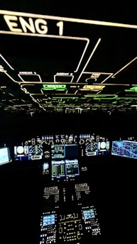 A cockpit filled with illuminated panels and displays showing various controls and instruments. The main displays are active with complex data and indicators. The environment is dark, highlighting the bright green, blue, and white lights from the instruments.