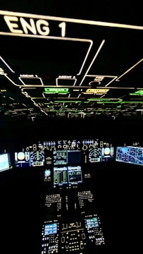 A cockpit filled with illuminated panels and displays showing various controls and instruments. The main displays are active with complex data and indicators. The environment is dark, highlighting the bright green, blue, and white lights from the instruments.