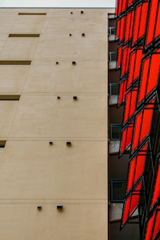 A modern architectural structure with a large beige wall, featuring minimalist black fixtures and rectangular windows. Adjacent to the wall, a series of vibrant red panels create a striking contrast, adding a dynamic element to the design.