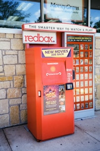 A Redbox movie rental kiosk is situated outside, with promotional signs indicating new movie releases. The kiosk is painted in red and has a keypad for user interaction. A stone wall forms the background, and there's signage above for related services.