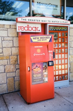 A Redbox movie rental kiosk is situated outside, with promotional signs indicating new movie releases. The kiosk is painted in red and has a keypad for user interaction. A stone wall forms the background, and there's signage above for related services.