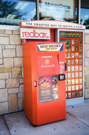 A Redbox movie rental kiosk is situated outside, with promotional signs indicating new movie releases. The kiosk is painted in red and has a keypad for user interaction. A stone wall forms the background, and there's signage above for related services.