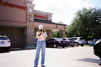 A young person stands in a parking lot in front of a pharmacy, wearing jeans, a tie-dye shirt, and a white cap, taking a photo or video with a smartphone. Several cars are parked, and there is a large tree providing greenery on the right side.