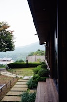 A serene outdoor setting featuring a modern building with a wooden deck overlooking a well-maintained garden. The garden includes manicured bushes, stone pathways, and a lush green lawn. In the background, rolling hills are visible under a partly cloudy sky, adding to the picturesque landscape.