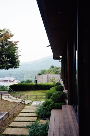 A serene outdoor setting featuring a modern building with a wooden deck overlooking a well-maintained garden. The garden includes manicured bushes, stone pathways, and a lush green lawn. In the background, rolling hills are visible under a partly cloudy sky, adding to the picturesque landscape.