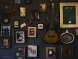 A dark-toned wall adorned with various framed art pieces and photographs, showcasing a diverse collection of portraits, artistic images, and a pair of playing cards. A guitar is hung centrally among these frames, adding a musical element, and an antique-style clock is also part of the collage. The arrangement appears artistic and vintage.