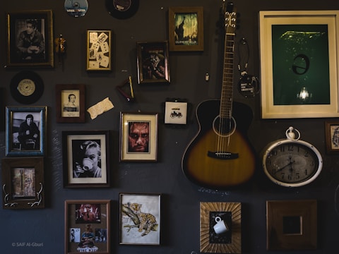 A dark-toned wall adorned with various framed art pieces and photographs, showcasing a diverse collection of portraits, artistic images, and a pair of playing cards. A guitar is hung centrally among these frames, adding a musical element, and an antique-style clock is also part of the collage. The arrangement appears artistic and vintage.