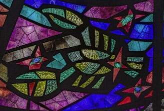 A colorful array of intricate stained glass fragments, featuring geometric shapes and patterns in vibrant hues. The pieces are textured and layered, creating a mosaic effect that captures and reflects light.