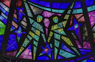 Colorful stained glass artwork featuring geometric patterns and the depiction of two human figures made from textured pieces of glass. The figures are composed of green and yellow hues, surrounded by a variety of blue, purple, and pink panels.