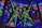 Colorful stained glass artwork featuring geometric patterns and the depiction of two human figures made from textured pieces of glass. The figures are composed of green and yellow hues, surrounded by a variety of blue, purple, and pink panels.