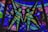 Colorful stained glass artwork featuring geometric patterns and the depiction of two human figures made from textured pieces of glass. The figures are composed of green and yellow hues, surrounded by a variety of blue, purple, and pink panels.