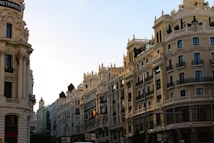 Elegant, historic buildings line a city street, bathed in warm, golden light from the setting sun. The architecture features ornate details with balconies and domed towers, creating a sense of grandeur. Flags adorn some of the buildings, adding a touch of color.