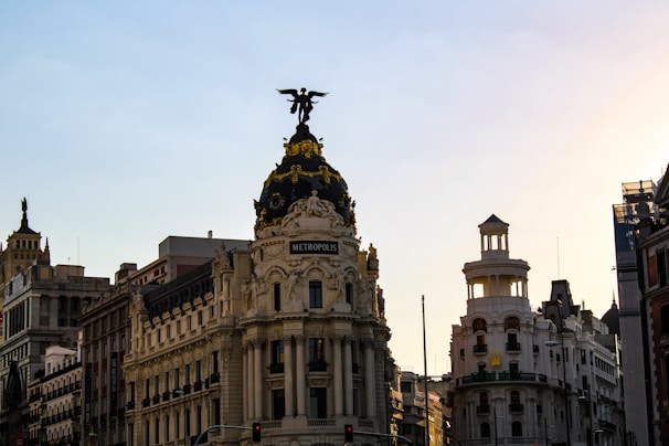 A beautifully ornate building with the name 'Metropolis' is the central focus. The building has a dark dome with a golden statue of a winged figure at the top. Surrounding the Metropolis building are other similarly styled buildings with detailed architectural designs. The sky is clear with a gradient from light blue to golden as the sun appears to be setting, casting a warm glow on the structures.