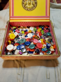 A box filled with a variety of colorful buttons, including different sizes and shapes. The box has a decorative lid with stripes and a circular emblem.