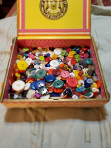 A box filled with a variety of colorful buttons, including different sizes and shapes. The box has a decorative lid with stripes and a circular emblem.