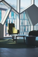 A modern interior space features angular geometric window designs with large panes allowing natural light to illuminate the room. In the foreground, there is a green upholstered armchair beside a round coffee table with a smaller side table nearby. The furnishings are placed on a green carpet that complements the chair, creating a cohesive and stylish setting.