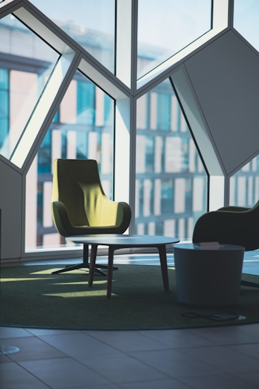 A modern interior space features angular geometric window designs with large panes allowing natural light to illuminate the room. In the foreground, there is a green upholstered armchair beside a round coffee table with a smaller side table nearby. The furnishings are placed on a green carpet that complements the chair, creating a cohesive and stylish setting.