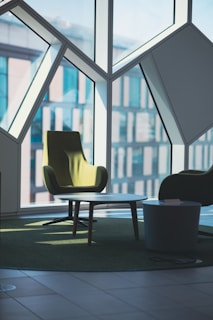 A modern interior space features angular geometric window designs with large panes allowing natural light to illuminate the room. In the foreground, there is a green upholstered armchair beside a round coffee table with a smaller side table nearby. The furnishings are placed on a green carpet that complements the chair, creating a cohesive and stylish setting.