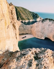 A secluded beach surrounded by steep, rugged cliffs with clear turquoise waters. A shipwreck is visible on the sandy beach, adding an element of mystery. The landscape is majestic and untouched, with a distant view of the sea meeting the horizon.