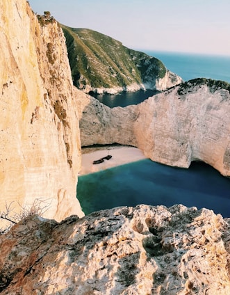 A secluded beach surrounded by steep, rugged cliffs with clear turquoise waters. A shipwreck is visible on the sandy beach, adding an element of mystery. The landscape is majestic and untouched, with a distant view of the sea meeting the horizon.