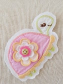 A colorful embroidered patch featuring a stylized turtle design with a pink shell adorned with layered fabric petals. A decorative button is placed at the center of the petals. The background is a textured, light-colored fabric.
