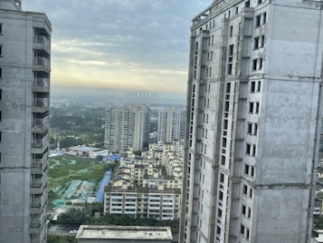 Tall, modern apartment buildings frame a distant view of a sprawling cityscape under a cloudy sky. The foreground features two prominent high-rise structures, while additional residential buildings are visible further in the background. A patch of green land is seen towards the bottom left.