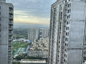 Tall, modern apartment buildings frame a distant view of a sprawling cityscape under a cloudy sky. The foreground features two prominent high-rise structures, while additional residential buildings are visible further in the background. A patch of green land is seen towards the bottom left.