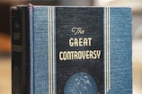 A hardcover book with a textured blue cover features the title 'The Great Controversy' printed in prominent gold letters. Vertical gold lines frame the title, adding an elegant touch. A circular embossed design appears near the bottom of the cover.
