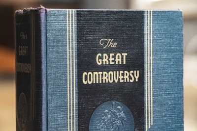 A hardcover book with a textured blue cover features the title 'The Great Controversy' printed in prominent gold letters. Vertical gold lines frame the title, adding an elegant touch. A circular embossed design appears near the bottom of the cover.