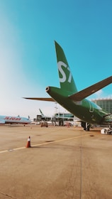 An airport scene with a green airplane bearing the number 'S7' parked on the runway. Another airplane with 'Jet2' written on it is visible in the background. The airport terminal is in view, along with several traffic cones and ground service equipment scattered across the tarmac.