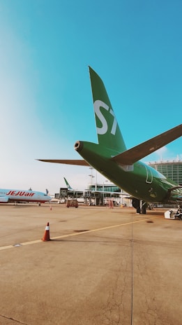 An airport scene with a green airplane bearing the number 'S7' parked on the runway. Another airplane with 'Jet2' written on it is visible in the background. The airport terminal is in view, along with several traffic cones and ground service equipment scattered across the tarmac.
