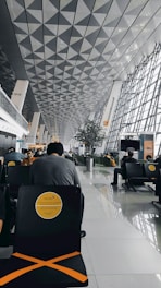 A modern airport terminal with a high, geometrically patterned ceiling and large glass windows. People are sitting in black chairs with yellow markings, and the area looks spacious and organized.