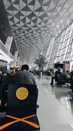 A modern airport terminal with a high, geometrically patterned ceiling and large glass windows. People are sitting in black chairs with yellow markings, and the area looks spacious and organized.