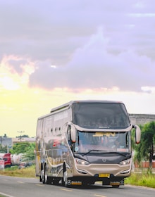 A large, modern bus with sleek, aerodynamic design is traveling on a road. The bus is branded with the Mercedes logo and has reflective windows. The road is surrounded by green foliage and some industrial buildings in the background. The sky is cloudy with patches of soft sunlight.