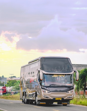 A large, modern bus with sleek, aerodynamic design is traveling on a road. The bus is branded with the Mercedes logo and has reflective windows. The road is surrounded by green foliage and some industrial buildings in the background. The sky is cloudy with patches of soft sunlight.
