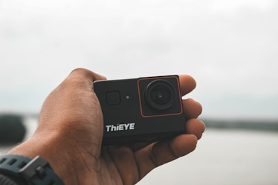 A hand holds a compact action camera close to the lens. The camera is labeled 'ThiEYE' and features a lens and power button. The background is blurred, indicating an outdoor setting.