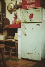 A vintage kitchen setting featuring a worn, pastel green refrigerator adorned with nostalgic stickers. On top of the fridge, red Coca-Cola crates are stacked. The adjacent wall showcases hanging kitchen utensils and decorative items. Below the counter, wooden stools and rustic elements evoke a retro atmosphere.