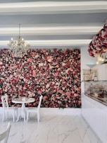A floral wall adorned with a dense arrangement of colorful flowers serves as the backdrop of a chic, modern café. A crystal chandelier hangs from the ceiling emitting a soft glow, while a small round table with white chairs sits in the foreground. The floor is made of a sleek, white marble-like material, and a pastry counter on the right displays an assortment of desserts under glass. The setting exudes elegance and sophistication.
