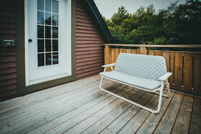 A cozy wooden deck features a simple white-framed bench with a quilted cushion. The background includes a wooden railing and a partially visible red and brown house. A glass-paneled door is on the left, and dense green foliage is visible beyond the railing.