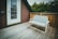 A cozy wooden deck features a simple white-framed bench with a quilted cushion. The background includes a wooden railing and a partially visible red and brown house. A glass-paneled door is on the left, and dense green foliage is visible beyond the railing.