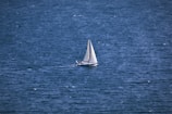 A lone sailboat with white sails navigates the expansive, deep blue sea. The water appears slightly choppy, indicating a breeze that may be propelling the vessel forward. The scene evokes freedom and solitude, with no other boats or land in sight.