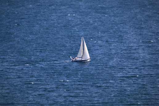 A lone sailboat with white sails navigates the expansive, deep blue sea. The water appears slightly choppy, indicating a breeze that may be propelling the vessel forward. The scene evokes freedom and solitude, with no other boats or land in sight.