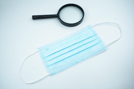 A black magnifying glass lies beside a light blue disposable face mask on a white surface.