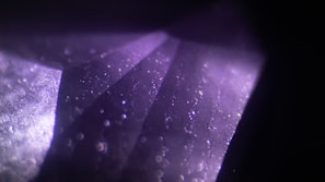 A close-up view of a geometric, crystalline structure with a purple hue. The surface is illuminated, revealing specks of reflected light and subtle textures resembling facets of a gem.
