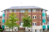 A red brick apartment building with multiple windows and a sloped roof. Two vibrant green trees are situated in front of the building, and the structure features teal-colored panels on the sides. The sidewalk leads up to the entrance, which is bordered by a black metal railing.
