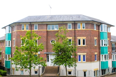 A red brick apartment building with multiple windows and a sloped roof. Two vibrant green trees are situated in front of the building, and the structure features teal-colored panels on the sides. The sidewalk leads up to the entrance, which is bordered by a black metal railing.